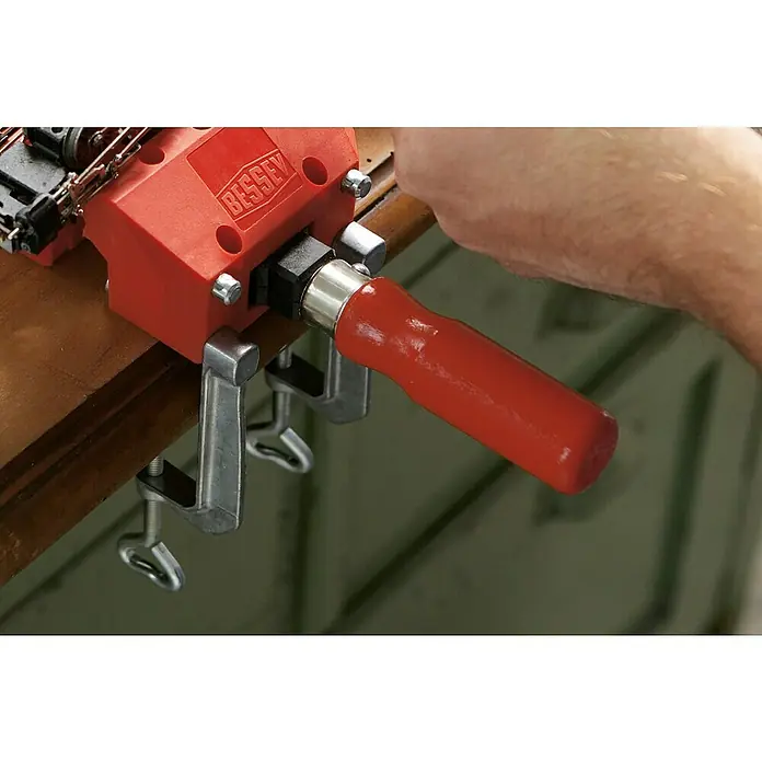 Bessey Tischklemme TK 6Spannweite: 60 mm - Eine rote Bessey- Schraubstock an einer Werkbank befestigt, mit einem silbernen Griff, der von einer Hand bedient wird.