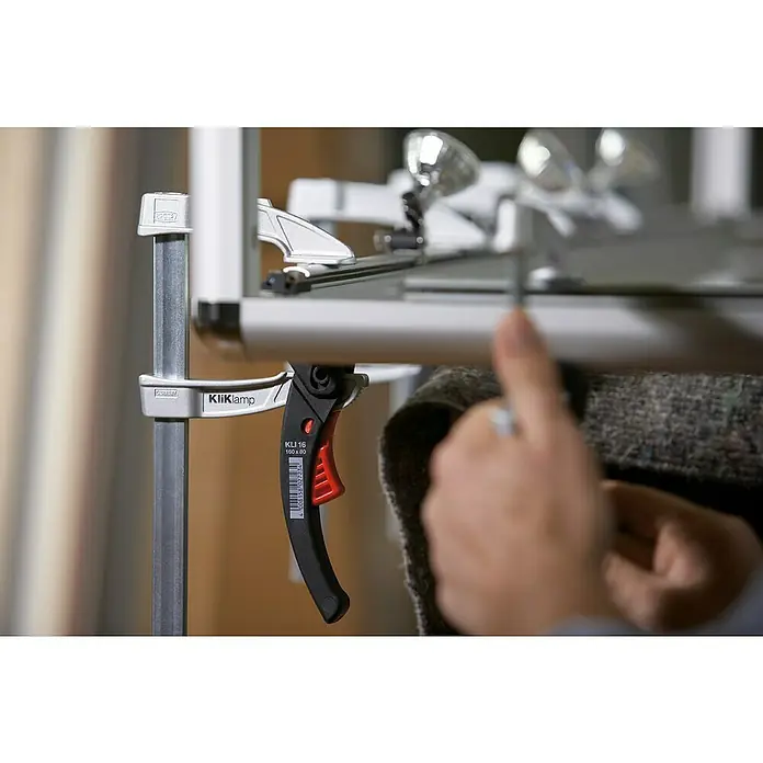Bessey Hebelzwinge KliKlamp KLI 16 - Spannweite: 160 mm, Spannkraft: 1.200 N, Ausladung: 80 mm - Nahaufnahme einer Bessey KliKlamp-Einhandzwinge mit einem schwarzen Griff und einem roten Auslöser, die an einer Metallstruktur befestigt ist.