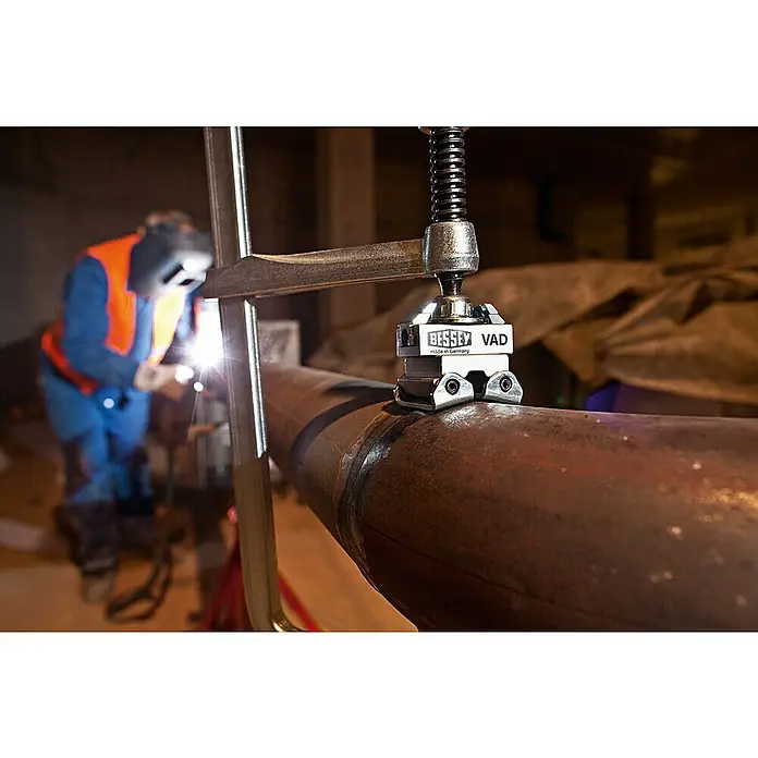 Bessey Spannaufsatz Vario VAD - Passend für: Bessey Tempergusszwingen, Bessey Ganzstahlzwingen GZ/GMZ - Bessey VAD Rohraufspannvorrichtung mit Schweißarbeiten im Hintergrund, ideal zum präzisen Fixieren von Rohren.