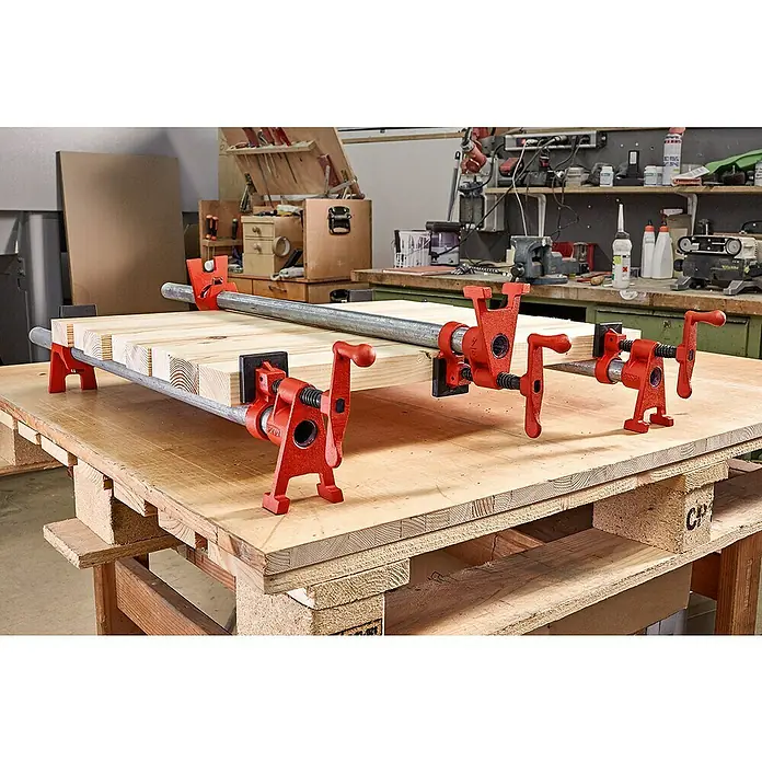 Bessey Rohr-Schraubzwinge BPC-H34 - Ausladung: 42 mm, Durchmesser Rohr: 19,05 mm - 26,9 mm - Eine Nahaufnahme von verleimten Holzbrettern, die mit roten Rohrklemmen auf einer Palette befestigt sind. Werkstatthintergrund ist sichtbar.