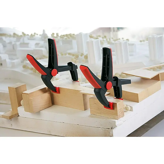 Bessey Federzwinge Clippix Vario XV 3-50 - Spannweite: 50 mm, Spannkraft: 80 N, Ausladung: 37 mm - Auf dem Bild sind zwei schwarze und rote Federklemmen zu sehen, die Holzstücke auf einem weißen Modell befestigen.