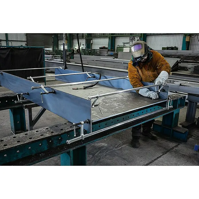 Bessey Zwingen-Set SLV 100 M - Spannweite: 1.000 mm, Spannkraft: 6.500 N, Ausladung: 120 mm - Schweißer bei der Arbeit an einer großen Metallkonstruktion, die mit Klammern auf einem Werkstisch befestigt ist. Schutz durch Helm und Handschuhe.