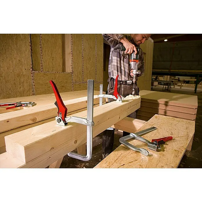 Bessey Hebelzwinge GH20 - Spannweite: 200 mm, Spannkraft: 8.500 N, Ausladung: 100 mm - Eine Werkstattaufnahme mit einem fokussierten Holzbearbeiter und einer robusten Schraubzwinge im Vordergrund.