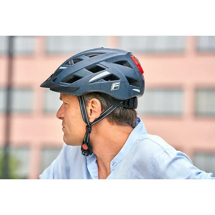 Fischer Fahrradhelm Urban Plus Brooklyn - S/M 52 - 59 cm, Schwarz - Ein grauer Fahrradhelm mit Belüftungsöffnungen und einem 'F'-Logo auf der Seite, getragen von einem Mann in einem hellblauen Hemd.