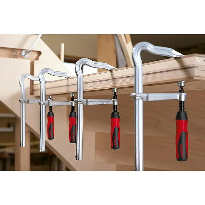 Bessey Schraubzwinge OMEGA GMZ - Spannweite: 250 mm, Spannkraft: 5.000 N, Ausladung: 120 mm - Mehrere BESSEY Ganzstahl-Schraubzwingen sind an einem Holzstück befestigt und fixieren es. Die Zwingen haben rote Griffe.