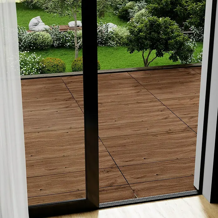 Marazzi Terrassenfliese Esterno 20T Woodliving Scuro - 40 x 120 x 2 cm, Braun, Matt - Nahaufnahme einer Holzterrasse, die durch eine offene Glasschiebetür mit schwarzem Rahmen einen Blick auf einen üppigen Garten freigibt.