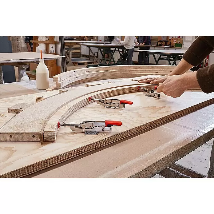 Bessey Schnellspanner STC-IHH25 - Spannweite: 16 mm, Spannkraft: 2.500 N - Hände befestigen gebogene Sperrholzschichten mit rot-griffigen Spannzwingen während eines Holzmontageprozesses.