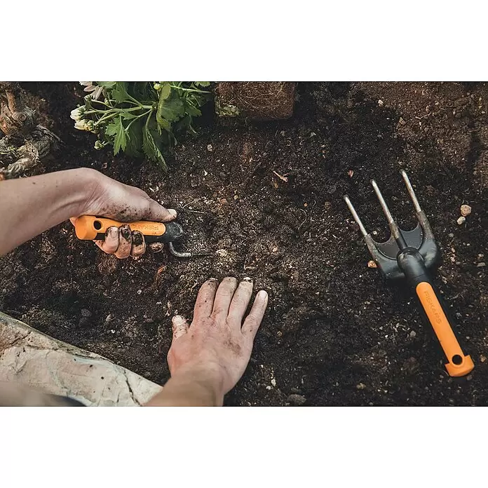 Fiskars Premium Pflanzkelle - Länge: 27,8 cm - Person gärtnert mit Fiskars Handkelle und Gabel mit orange-schwarzem Griff in dunkler Erde.