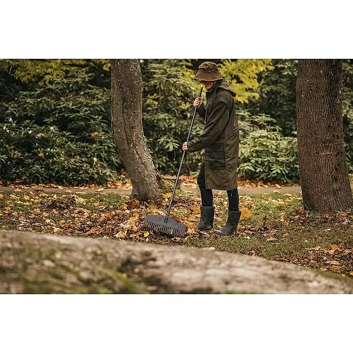 Fiskars Solid Laubbesen M - Arbeitsbreite: 41 cm - Person in langem Mantel und Hut benutzt eine schwarze, langstielige Laubharke, um Herbstlaub auf dem Rasen zu sammeln.