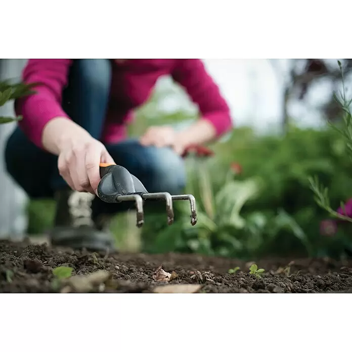 Fiskars Premium Kleinrechen - Länge: 25 cm, Stahl - Nahaufnahme einer Person, die eine schwarz-orange Harke zum Kultivieren von Gartenerde benutzt.