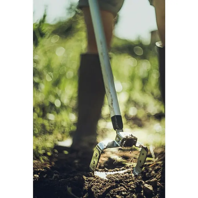 Fiskars Xact Jäter - Arbeitsbreite: 13,5 cm, Zweischneidige Klinge - Nahaufnahme einer Gartenhacke, die in die Erde gesteckt wird, um den Garten zu bearbeiten, wobei der Fokus auf dem Werkzeug und der umgebenden Erde liegt.