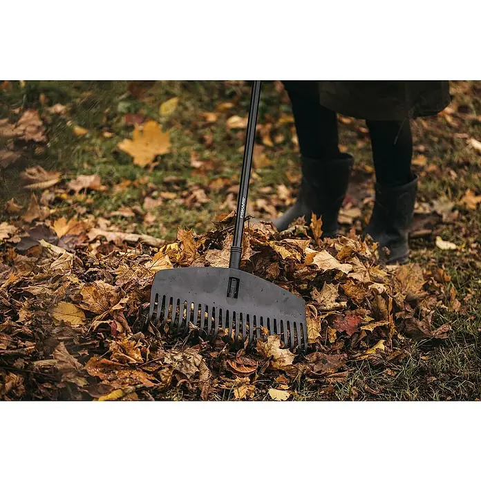 Fiskars Solid Laubbesen M - Arbeitsbreite: 41 cm - Nahaufnahme eines Fiskars Laubrechens, der in einem Haufen bunter Herbstblätter steckt, während eine Person im Hintergrund steht.
