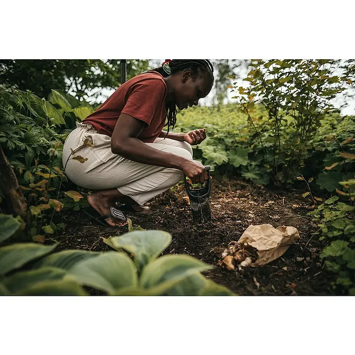 Fiskars Solid Hand-Blumenzwiebelpflanzer - 11,7 x 24,3 cm - Eine Frau hockt in einem Garten und pflanzt Blumenzwiebeln mit einem kleinen Spaten. Die braune Papiertüte mit den Zwiebeln liegt auf dem Boden.