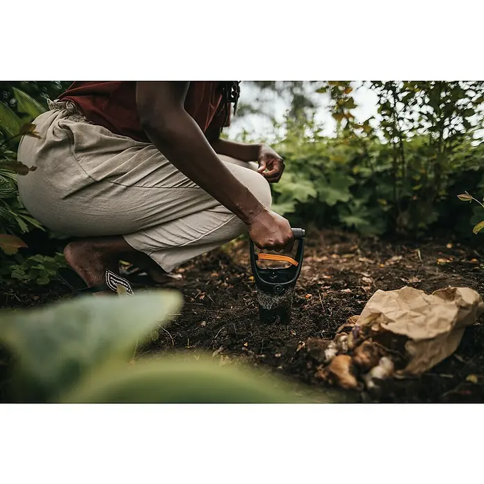 Fiskars Solid Hand-Blumenzwiebelpflanzer - 11,7 x 24,3 cm - Eine Person benutzt einen Pflanzholz, um Blumenzwiebeln in einem Garten zu pflanzen, mit einer Papiertüte voller Zwiebeln daneben.