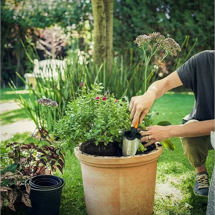 Fiskars Premium Blumenkelle - Länge: 27 cm, Stahl - Eine Person pflanzt Blumen mit einer silbernen Gartenschaufel in einen großen Terrakotta-Topf in einem sonnigen Garten.