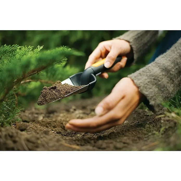 Fiskars Premium Blumenkelle - Länge: 27 cm, Stahl - Nahaufnahme einer Person, die mit einer kleinen Schaufel Erde in ein Pflanzloch gibt, um eine Pflanze einzusetzen.
