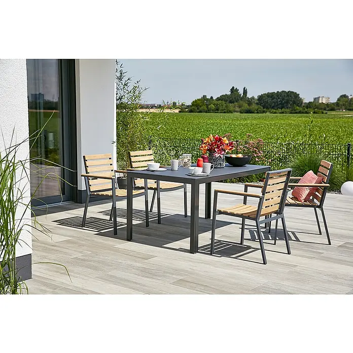 Gartenmöbel-Set Maja/Carina - 5 -tlg., Glas/Aluminium/Akazie/Stahl, Anthrazit/Naturbraun, Gartenstuhl stapelbar - Ein grauer Gartentisch mit vier passenden Holzstühlen mit Armlehnen auf einer Terrasse mit Blick auf ein Feld.