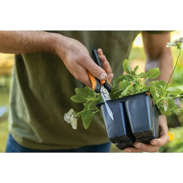 Fiskars Solid Mehrzweck-Schere SP320 - Länge: 20,6 cm - Nahaufnahme einer Person, die eine schwarze Pflanzschale mit einer orangefarbenen Gartenschere aufschneidet, um die Pflanze umzutopfen.