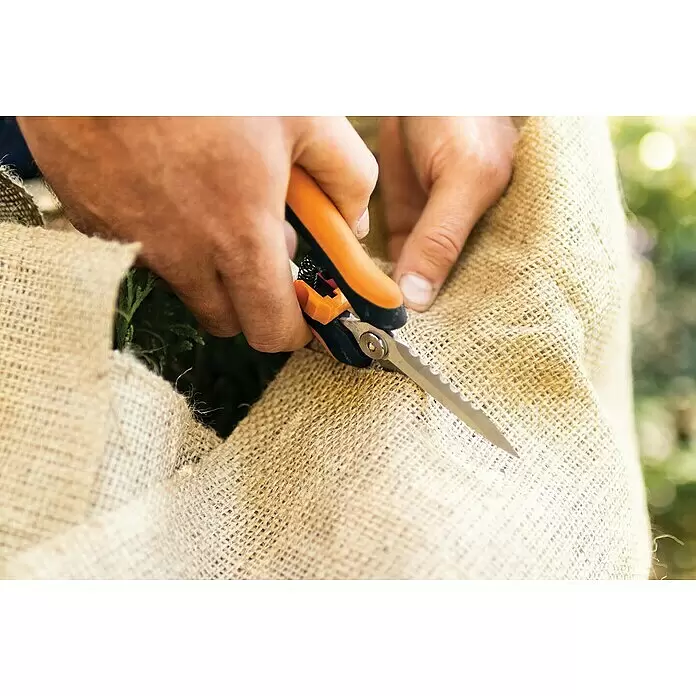 Fiskars Solid Mehrzweck-Schere SP320 - Länge: 20,6 cm - Nahaufnahme von Händen, die orange-schwarze Gartenschere mit gezackter Klinge verwenden, um Sackleinen um eine Pflanze zu schneiden.