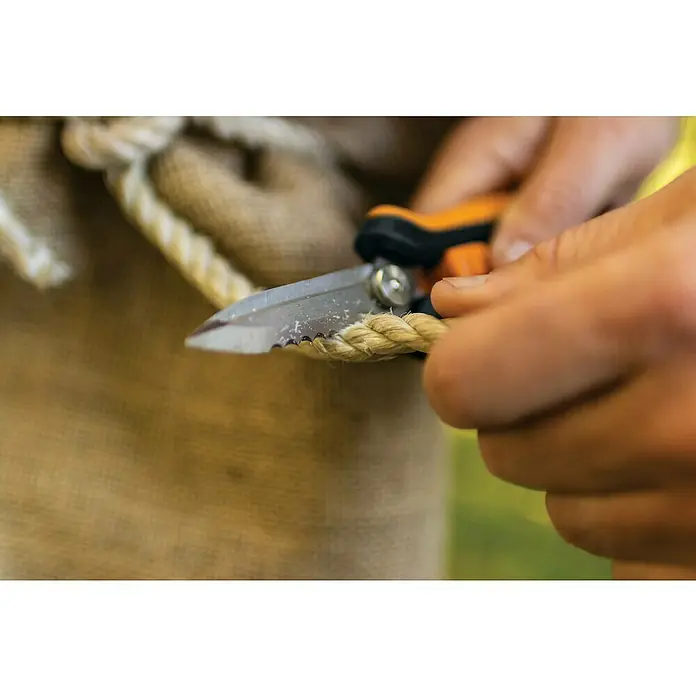 Fiskars Solid Mehrzweck-Schere SP320 - Länge: 20,6 cm - Nahaufnahme von Händen, die ein Seil mit einer kleinen Schere mit orangefarbenen Griffen durchschneiden, wobei der Fokus auf dem Schneidvorgang liegt.