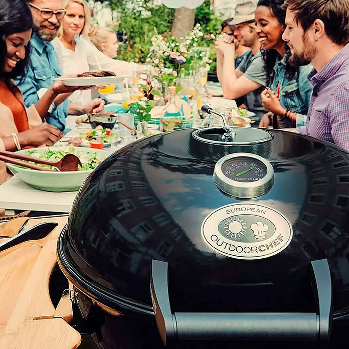 Outdoorchef Kugelgrill CHELSEA 480 C - Durchmesser Grillfläche: 45 cm, Schwarz - Draufsicht auf einen schwarzen Outdoorchef-Kugelgrill mit Temperaturanzeige und dem Logo vor einer geselligen Grillparty.