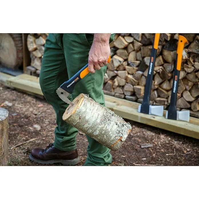 Fiskars WoodXpert Sappie XA2 - 35 cm - Nahaufnahme einer Person mit grüner Hose, die eine Fiskars XA20 Axt hält, in die ein Holzstück eingekeilt ist. Holzscheite im Hintergrund.