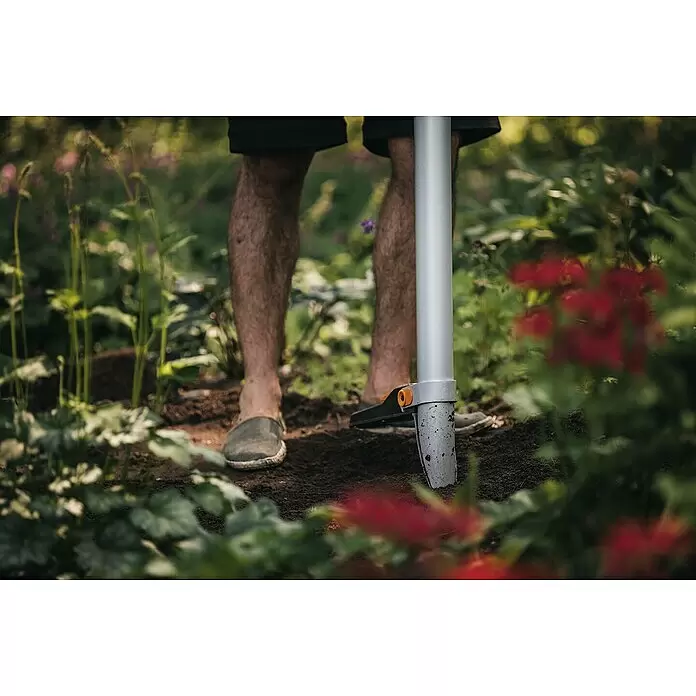 Fiskars Xact Hand-Blumenzwiebelpflanzer - Länge: 105 cm - Eine Person benutzt ein silbernes Gartengerät mit orangefarbenem Akzent und drückt es in die dunkle Erde im Garten.