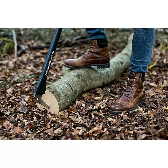 Fiskars WoodXpert Sappie XA22 - Länge: 78 cm - Schwarze Fiskars XA22 Axtklinge in einen gefällten Holzstamm geschlagen, umgeben von braunen Stiefeln und blauen Jeans auf Laub.