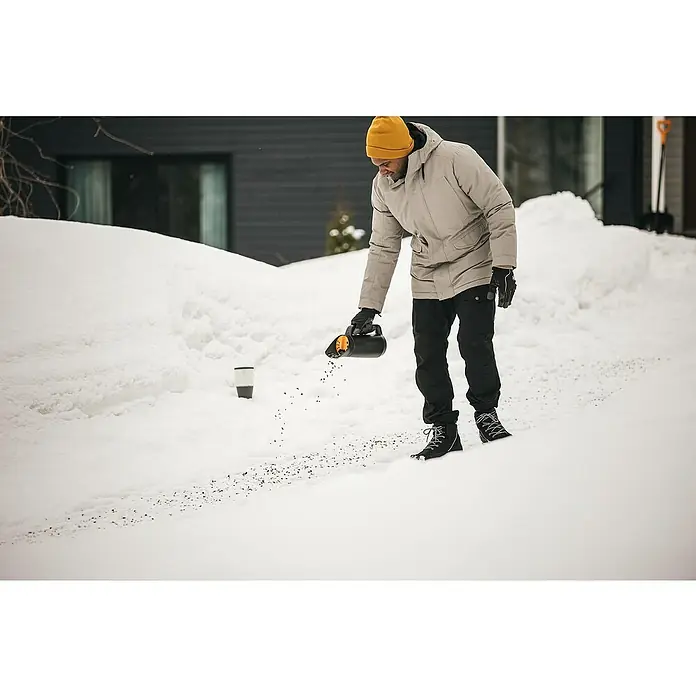 Fiskars Solid Handstreuer - 2 l - Mann streut mit einem manuellen Streuer Salz auf einen verschneiten Weg, um die Oberfläche sicherer zu machen. Gelbe Mütze und Winterjacke.