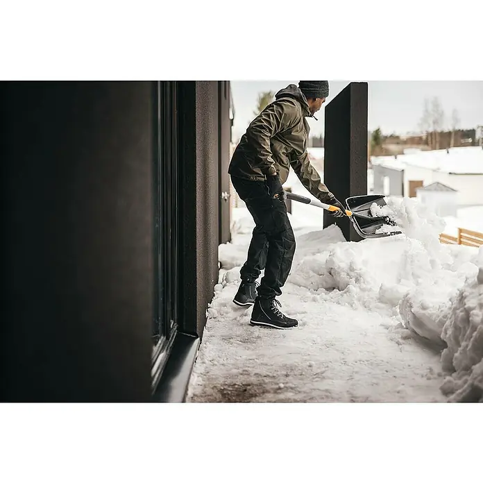 Fiskars X-series Schneeschaufel - Breite: 36,2 cm, Teleskopstiel - Ein Mann mit einer Fiskars Schneeschaufel räumt Schnee vor einem modernen Haus, bekleidet mit Winterkleidung für die Kälte.
