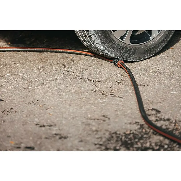 Fiskars FiberComp Reparator - Passend für: 3/4″-Schläuche Ø 19 mm - Ein schwarzer Gartenschlauch mit orangen Streifen liegt unter einem Reifen auf einer Betonoberfläche.