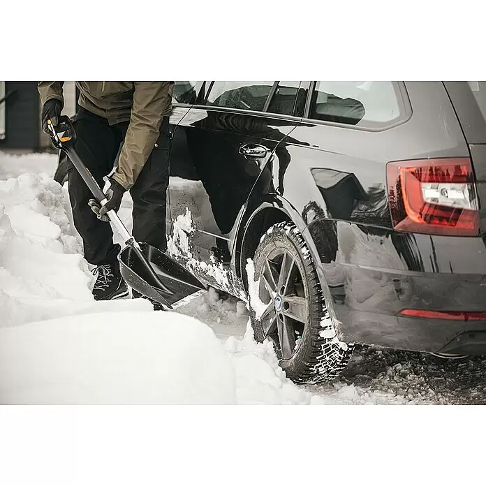 Fiskars X-series Schneeschaufel - Breite: 29 cm, Teleskopstiel - Eine Person benutzt eine schwarz-graue Schneeschaufel, um Schnee neben einem Auto zu räumen.