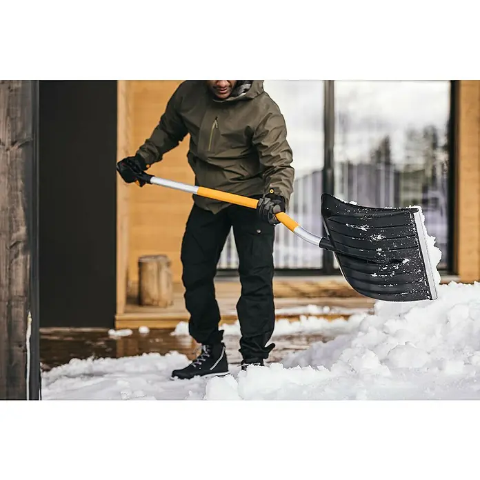 Fiskars X-series Schneeschieber - Breite: 53 cm, Ergostiel - Ein Mann schaufelt mit einer schwarzen Fiskars Schneeschaufel Schnee vor einem Haus. Die Schaufel hat einen gelben Griff.