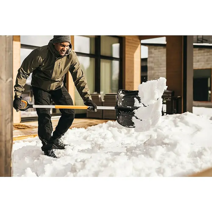 Fiskars X-series Schneeschieber mit Aufbug - Breite: 53 cm - Ein Mann schaufelt Schnee mit einer Fiskars Schneeschaufel vor einem modernen Haus.
