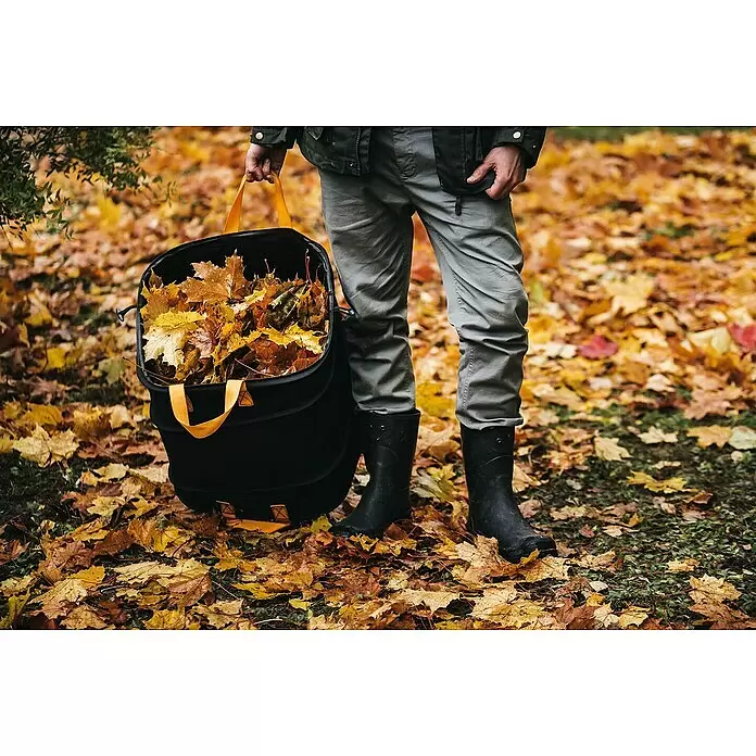 Fiskars Ergo Pop-Up Gartenabfallsack - 175 l - Person trägt eine schwarze, zusammenklappbare Laubsammeltasche mit orangefarbenen Griffen, gefüllt mit Herbstblättern.