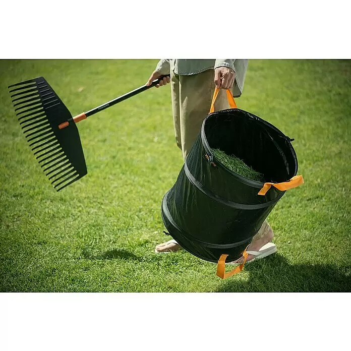 Fiskars Solid Pop-Up Gartenabfallsack - 172 l - Eine Person nutzt eine schwarze Harke und einen schwarzen Pop-up-Gartensack, gefüllt mit grünem Grasschnitt, auf einem Rasen.