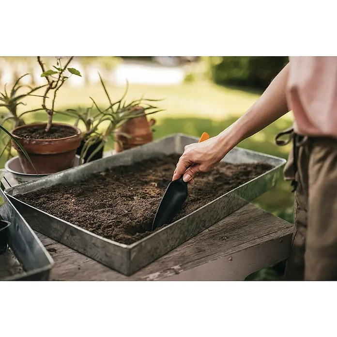 Fiskars Solid Pflanzkelle Saatgut - 29,8 cm - Nahaufnahme einer Person, die mit einer schwarzen Gartenschaufel Erde in einem metallenen Pflanzgefäß umgräbt, um Pflanzen einzusetzen.