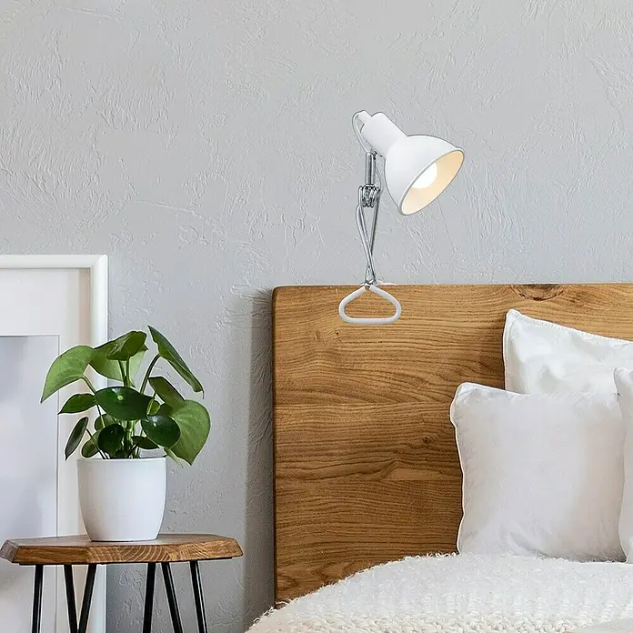Briloner Klemmleuchte Fix - 8 W, L x B x H: 30,5 mm x 11 cm x 13 cm, Farbe Gehäuse: Weiß - Nahaufnahme einer weißen Klemmlampe, die an einem Holzbett befestigt ist, mit einem Nachttisch mit einer Pflanze daneben.
