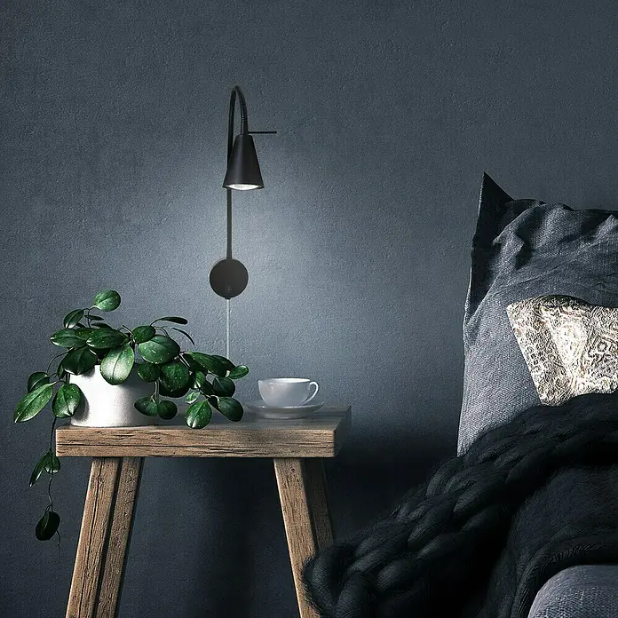 Briloner LED-Wandstrahler Tusa - 4 W, L x B x H: 5,8 x 20,5 x 45 cm, Schwarz, Warmweiß - Elegante schwarze Wandleuchte über einem Nachttisch mit Pflanze und Tasse, die eine gemütliche und stilvolle Atmosphäre schafft.