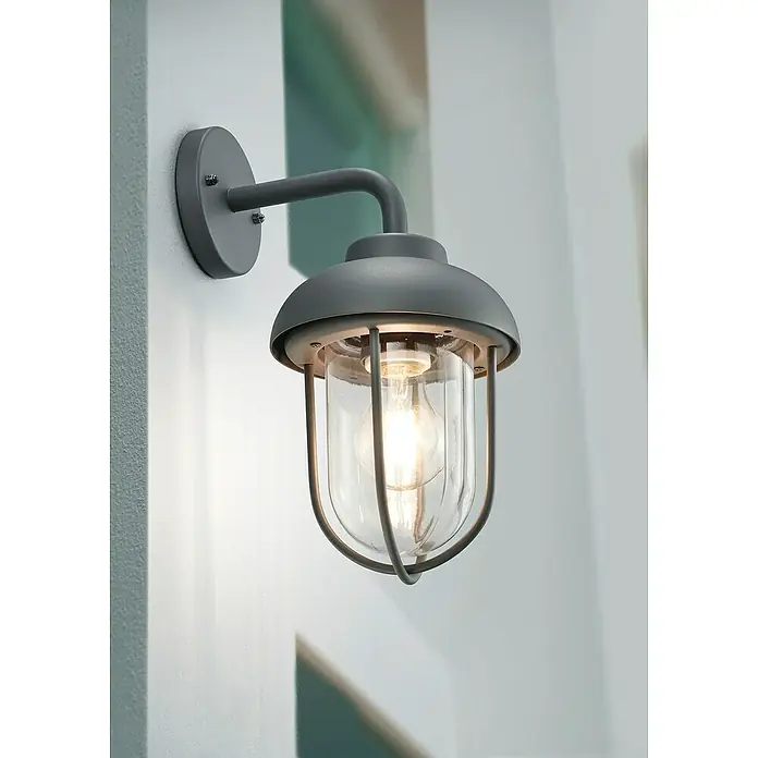 Trio Leuchten Außenwandleuchte Duero - 40 W, L x B x H: 21,3 x 16 x 33 cm, Anthrazit, IP44 - Nahaufnahme einer grauen Wandleuchte im Außenbereich mit klarem Lampenschirm und dekorativem Käfig, die ein warmes Licht ausstrahlt.