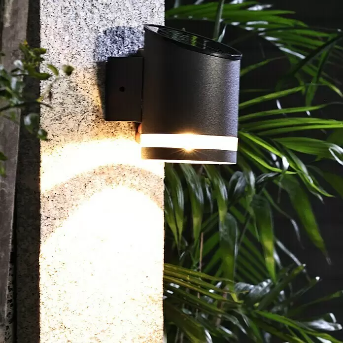 BAUHAUS Solar-Außenwandleuchte - 0,1 W, L x B x H: 12 x 9 x 12,5 cm, Anthrazit, 15 lm - Schwarze zylindrische Solar-Wandlampe beleuchtet eine Steinwand. Mit Leuchtband und Solarpanel oben.