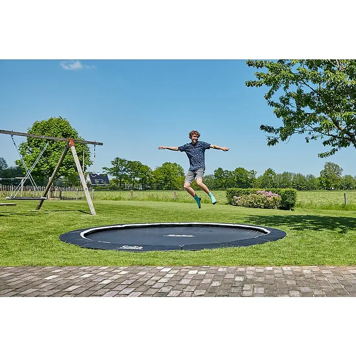 Salta Trampolin Royal Baseground - Durchmesser: 396 cm, Schwarz - Ein junger Mann springt auf einem runden Salta-Trampolin im Garten mit grüner Rasenfläche.