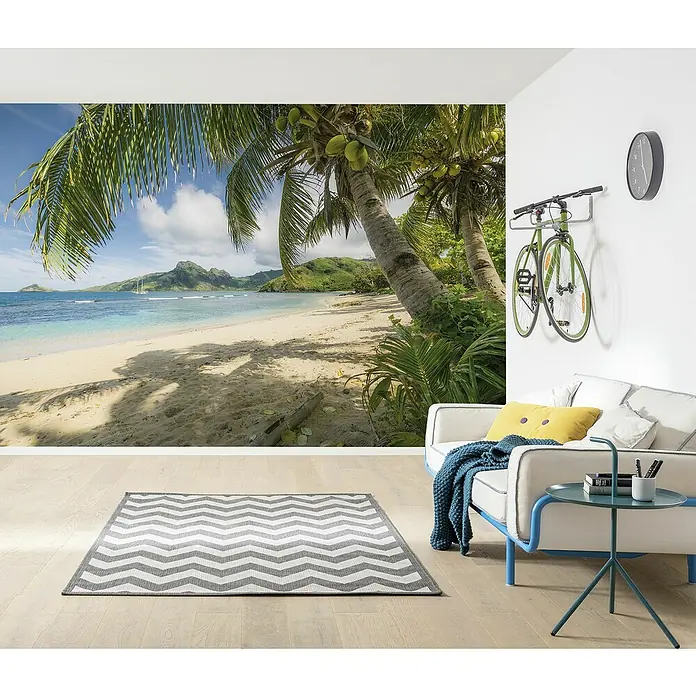 Komar Colours Fototapete Castaway - B x H: 368 x 254 cm, Papier - Wohnzimmer mit gemustertem Teppich, Sofa, Beistelltisch, Wanddekoration und Fahrradaufhängung. Helle und moderne Einrichtung mit tropischem Strandmotiv.