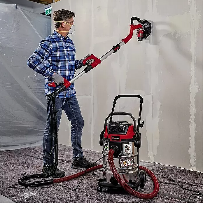 Einhell Expert Elektro-Nass- & Trockensauger TE-VC 2230 SACL - 1.400 W, 30 l - Mann schleift Wand mit rotem/silbernem Langhalsschleifer, verbunden mit rotem/verchromtem Nass-Trockensauger zur Staubabsaugung.