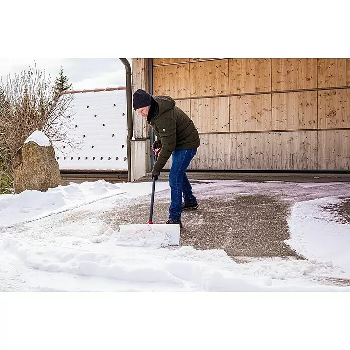 Freund Victoria Schneeschieber Snow pusher - Breite: 61 cm, Langstiel - Ein Mann schaufelt Schnee mit einer weiß-schwarzen Schneeschaufel mit rotem Griff auf einer Einfahrt.