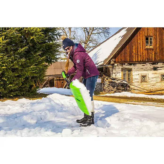 Freund Victoria Kunststoff-Schneeschaufel - Breite: 40 cm, Material Stiel: Aluminium - Eine Frau schaufelt Schnee mit einer grünen Schneeschaufel vor einem rustikalen Haus. Sie trägt eine warme Jacke und eine Mütze.