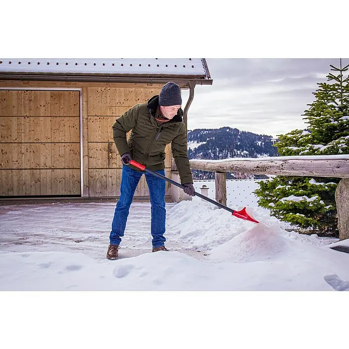 Freund Victoria Schneeschieber Snow pusher - Breite: 61 cm, Langstiel - Ein Mann in Winterkleidung schaufelt mit einer roten Schneeschaufel Schnee vor einem Haus mit schneebedeckten Bergen im Hintergrund.