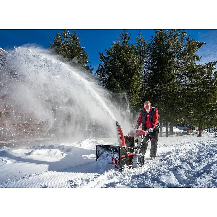 AL-KO Premium Benzin-Schneefräse SnowLine 620 E III - 6,2 kW, 302 cm³, Arbeitsbreite: 62 cm - Ein Mann benutzt eine AL-KO Schneefräse, um Schnee von einer Auffahrt zu räumen, wobei der Schnee hoch in die Luft geschleudert wird.