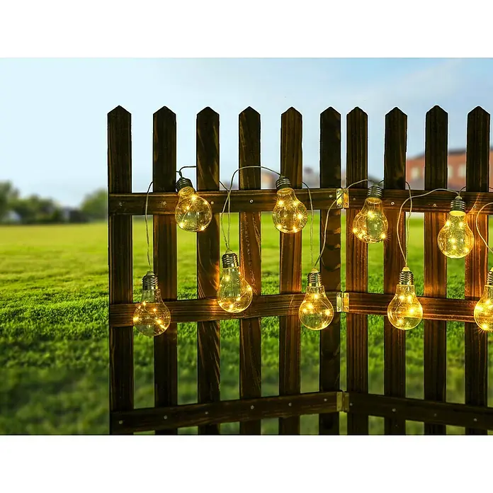 Globo Solar-Lichterkette - Klar - Eine Lichterkette mit Edison-Glühbirnen, die an einem Holzzaun befestigt ist, beleuchtet den Garten mit warmem Licht und schafft eine gemütliche Atmosphäre.