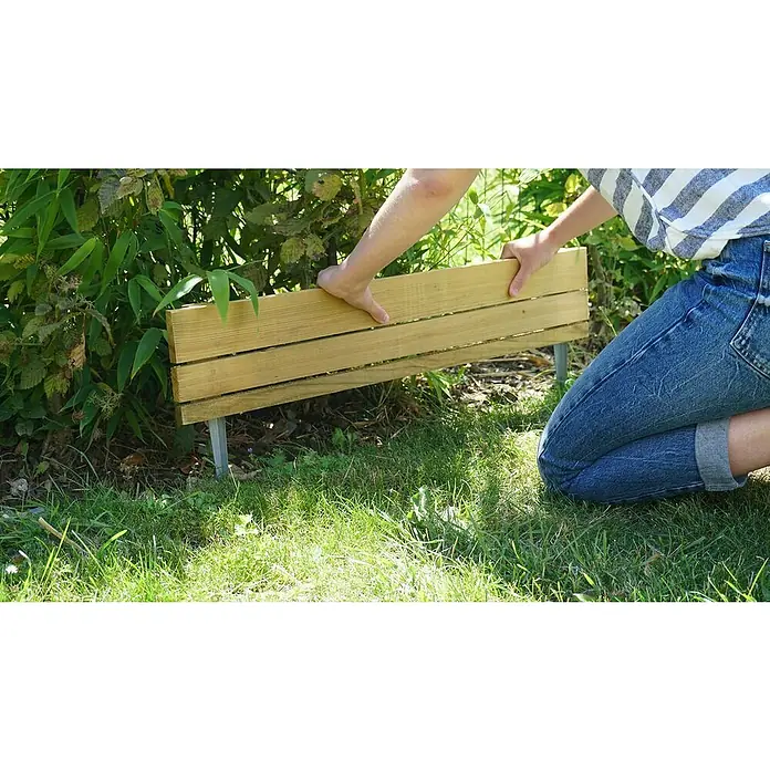 Cerland Bordura Hakea - L x An x Al: 30 x 3,9 x 80 cm, Madera, Natural - Una persona se arrodilla en el césped, sujetando un borde de jardín de madera con estacas de metal para crear un borde limpio para un parterre.